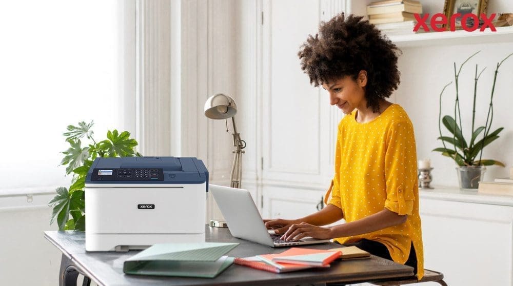 Een persoon met krullend haar zit glimlachend op een laptop te typen aan een bureau, met een compacte Xerox C310-printer naast de laptop. Dit is een voorbeeld van een moderne thuiskantooropstelling voor productiviteit.
