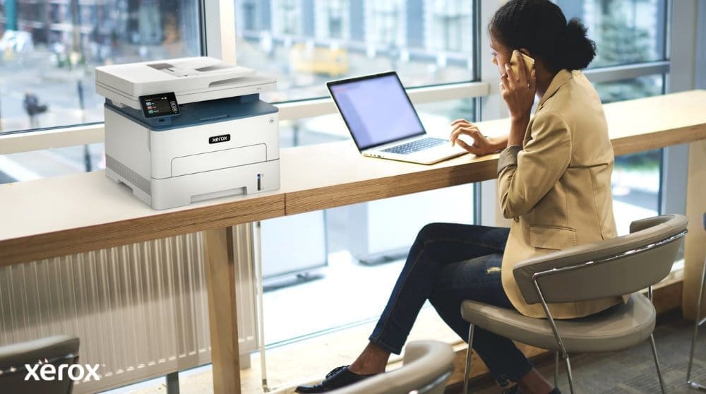 Xerox® Print en Scan Experience: handig en verbonden Iemand zit aan een hoge tafel bij een groot raam te telefoneren en op een laptop te werken. Een Xerox B235DNI multifunctionele printer staat handig naast hem op tafel, wat gebruiksgemak in een moderne werkomgeving suggereert.
