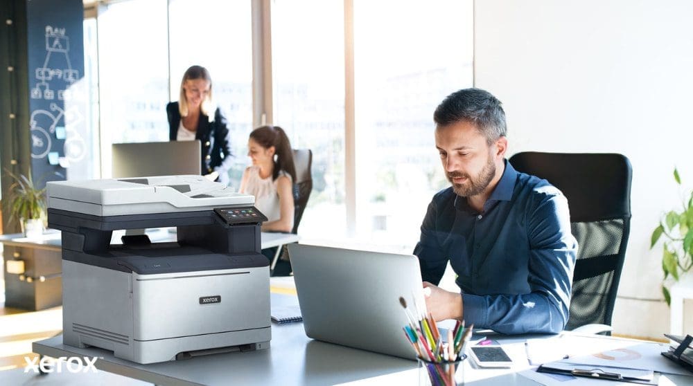 Une personne est assise à un bureau, les yeux rivés sur un ordinateur portable avec une imprimante multifonction Xerox C325 à côté. En arrière-plan, deux autres personnes travaillent également à leur bureau, dans un environnement lumineux.