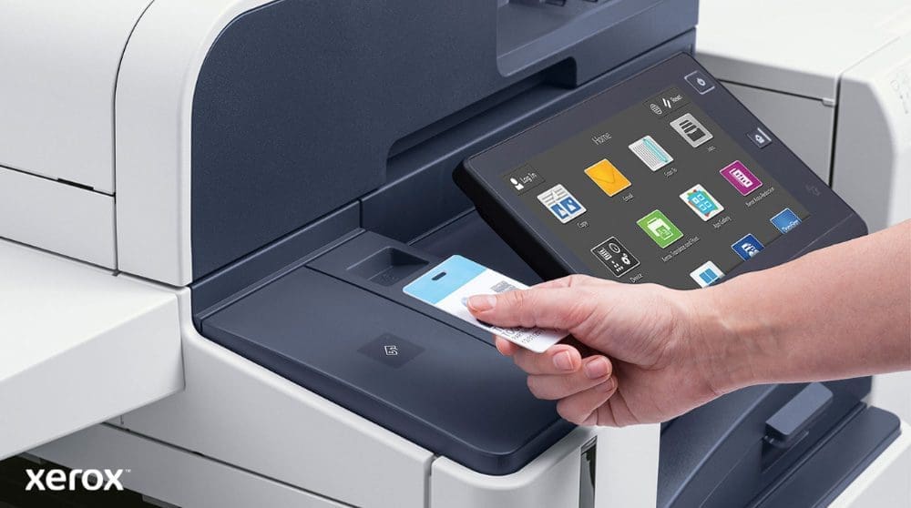 Xerox® Workplace Cloud en Suite: Veilige authenticatie en slimme interface Een hand houdt een beveiligingspas vast en scant deze bij de kaartlezer van een moderne multifunctionele Xerox-printer met een groot touchscreen-display waarop verschillende toepassingspictogrammen worden weergegeven.