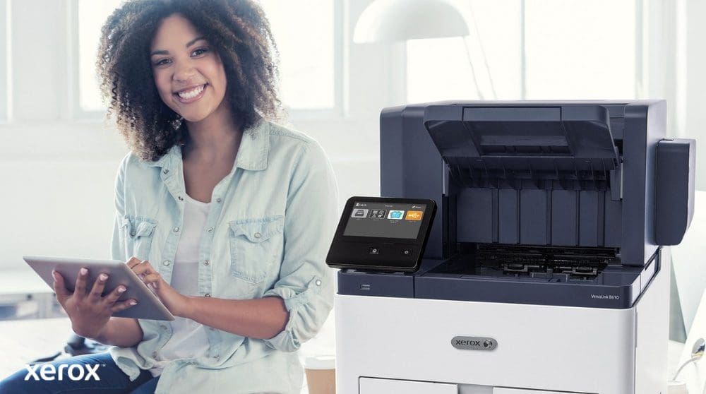 Xerox® Workplace Cloud en Suite: gebruiksvriendelijke integratie Een glimlachende persoon zit naast een witte Xerox VersaLink B610-printer, houdt een tablet vast en kijkt naar de camera. Dit benadrukt het gebruiksvriendelijke karakter van de Xerox-oplossingen.